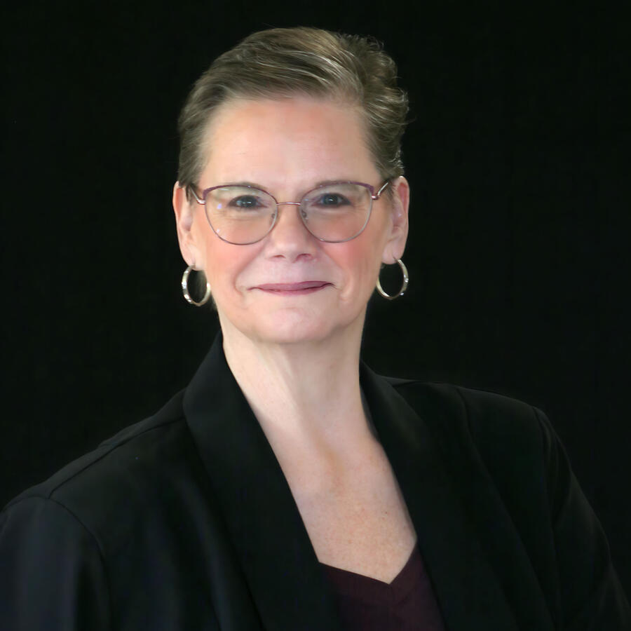 Jerri Glover Headshot of Jerri Glover. A smiling woman with short, dark hair, hoop earrings, and glasses. She is wearing business attire. The background is a flat, black color.